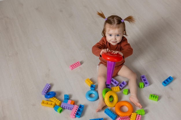 cheerful baby plays with a multicolored pyramid and other educational toys sitting on the floor