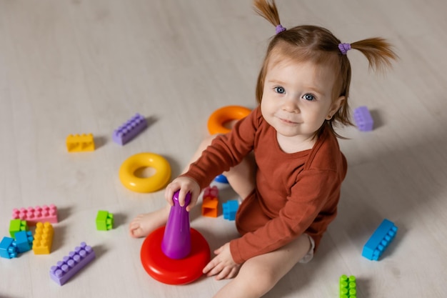 cheerful baby plays with a multicolored pyramid and other educational toys sitting on the floor
