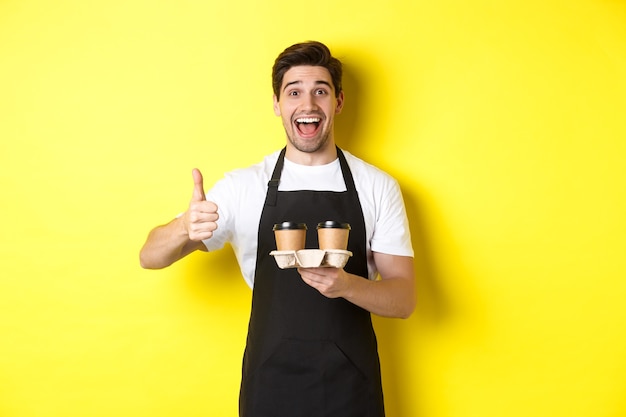 Cheerful barista recommend his cafe, holding takeaway coffee cups and showing thumbs up, standing against yellow wall