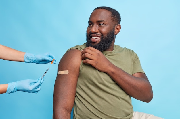 Photo cheerful bearded adult afro american man got covid 19 vaccine looks with happy expression at nurse shows shoulder with adhesive tap poses against blue wall