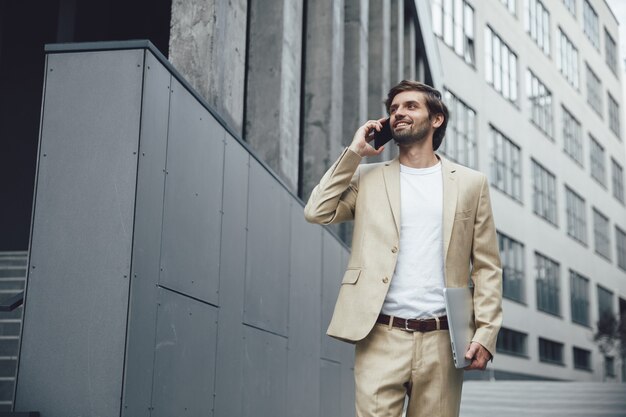 Веселый бородатый бизнесмен разговаривает по смартфону, стоя на улице с портативным ноутбуком