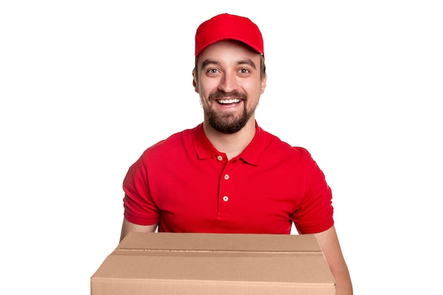 Cheerful bearded delivery man in red uniform and cap looking with friendly smile while carrying big cardboard box