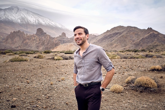 Cheerful bearded handsome male standing on dry grassy terrain near amazing mountainous in cloudy day
