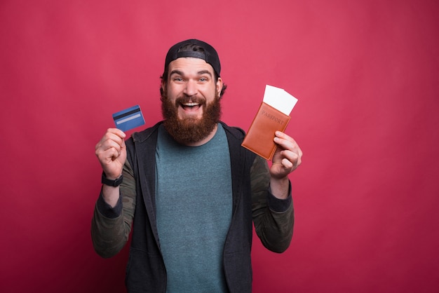 Cheerful bearded hipster is holding his passport and credit card on red pink