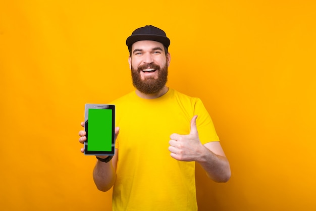 Cheerful bearded hipster man showing green screen on tablet and thumb up