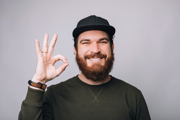 Cheerful bearded hipster man showing OK gesture over white wall