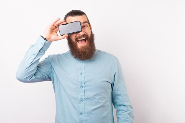 Uomo barbuto allegro in casual tenendo lo smartphone sugli occhi e sorridente