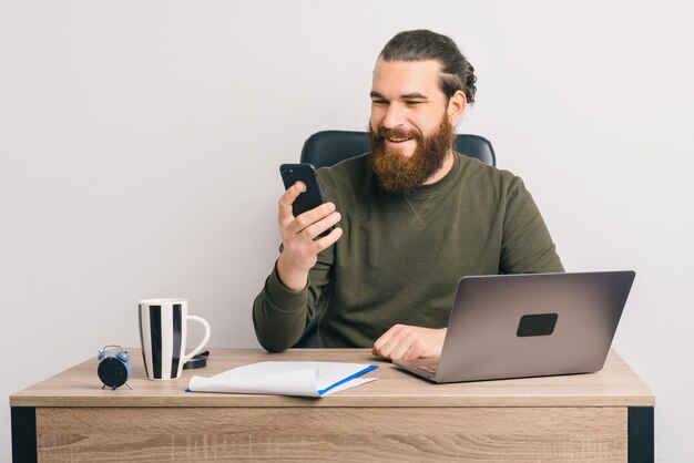 Cheerful bearded man is smiling while he is using his phone and laptop.