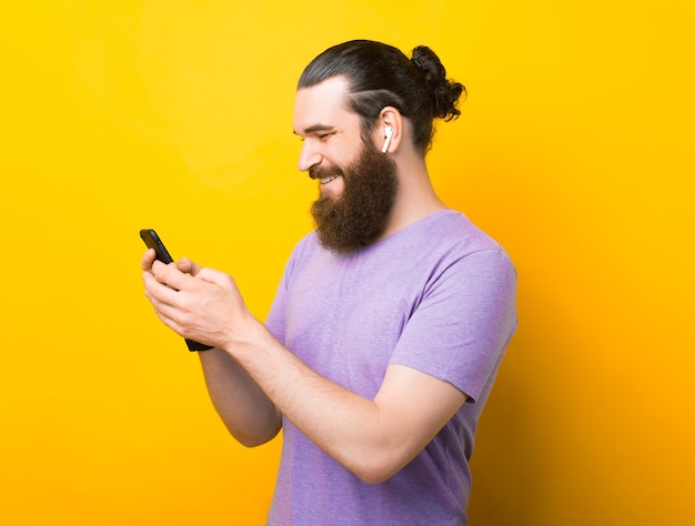 Cheerful bearded man is typing on his phone a message while listening to the music in ear pods.