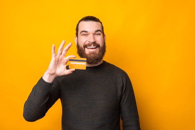 Cheerful bearded man smiling and showing credit card over yellow