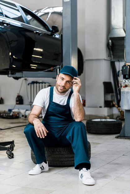 Cheerful bearded man touching cap and sitting on car tire