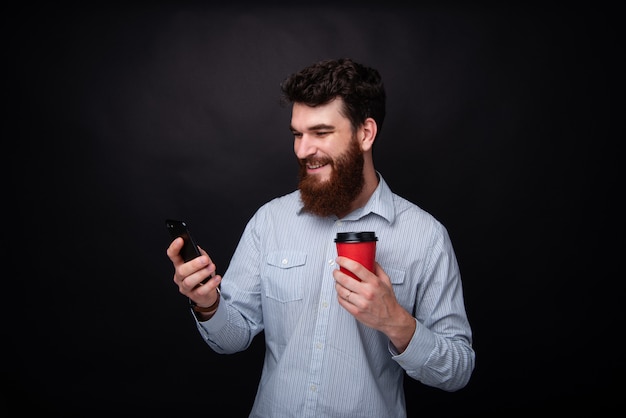 暗い孤立した背景の上に一杯のコーヒーを押しながらメッセージを入力する陽気なひげを生やした男