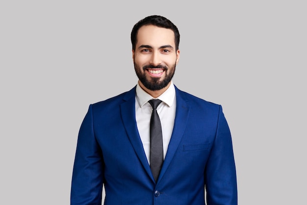 Cheerful bearded man with charming smile, standing and looking at camera, expressing positive emotions, wearing official style suit. Indoor studio shot isolated on gray background.