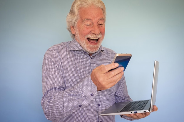 Cheerful bearded senior man holding laptop while using mobile phone 70 years old man laughing in video chat enjoying technology and social