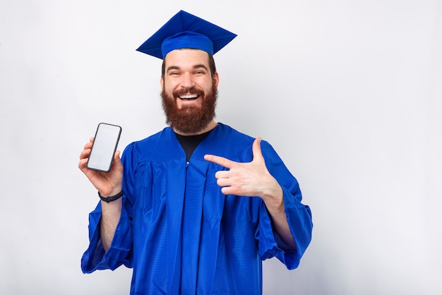 Cheerful bearded student man in blue bachelor pointing at smartphone