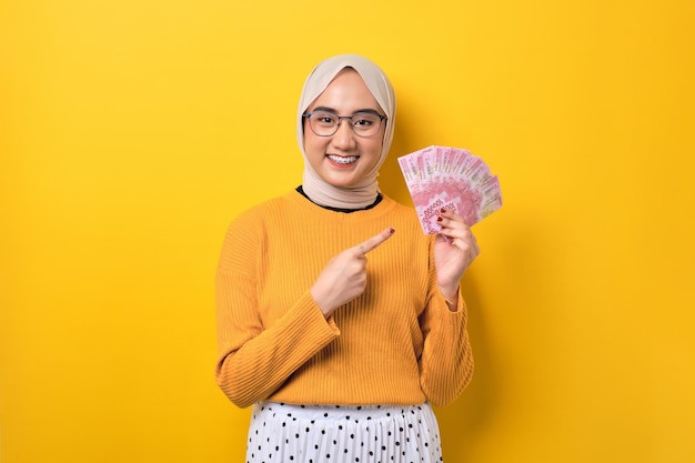 Cheerful beautiful Asian girl wearing hijab pointing finger at money banknotes isolated on yellow background