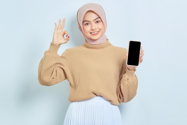 Cheerful beautiful Asian muslim woman in brown sweater and hijab showing blank screen mobile phone showing ok sign isolated over white background