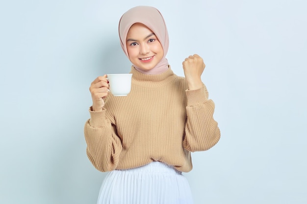 갈색 스웨터를 입은 쾌활한 아름다운 아시아 이슬람 여성이 커피잔을 들고 흰색 배경에 격리된 행운을 축하합니다.