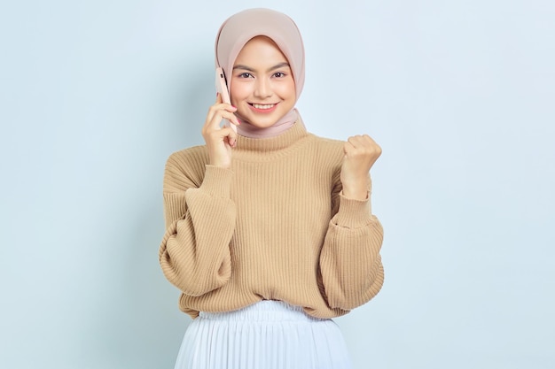 Cheerful beautiful Asian muslim woman in brown sweater talking mobile phone while rejoicing luck isolated over white background People religious lifestyle concept