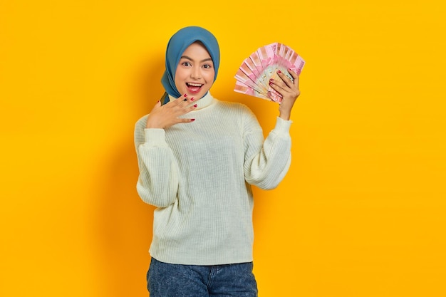 Cheerful beautiful Asian Muslim woman in white sweater holding cash money in Indonesian rupiah banknotes isolated over yellow background ​People religious lifestyle concept