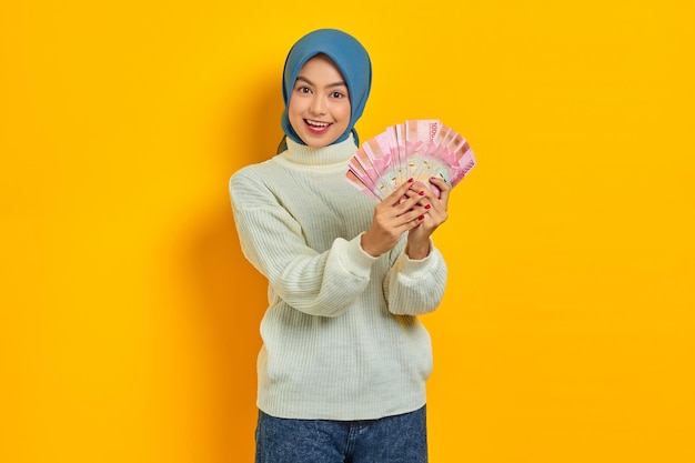 Cheerful beautiful asian muslim woman in white sweater holding\
fan of cash in rupiah banknotes isolated on yellow background\
people religious lifestyle concept