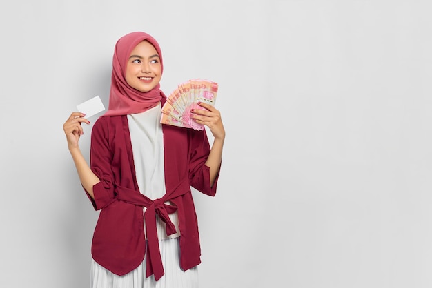Cheerful beautiful asian woman in casual shirt and hijab\
holding indonesian rupiah banknotes, showing credit card isolated\
over white background. people religious lifestyle concept