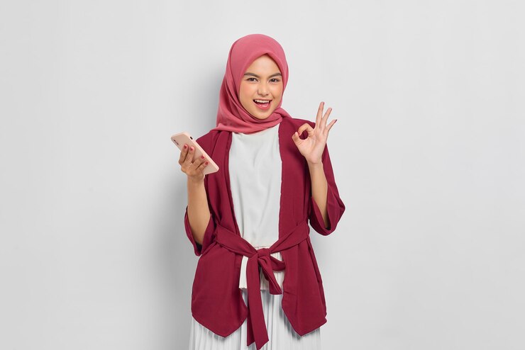 カジュアルなシャツとヒジャーブの陽気な美しいアジアの女性は、携帯電話を保持し、白い背景の上に分離されたokサインを身振りで示します。人々の宗教的なライフスタイルの概念