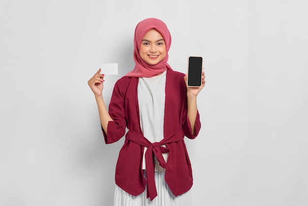 Cheerful beautiful asian woman in casual shirt and hijab holding a mobile phone with a blank screen, showing credit card isolated over white background. people religious lifestyle concept