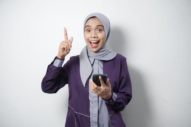 Cheerful beautiful Asian woman in casual shirt and hijab using mobile phone pointing empty space isolated by white background