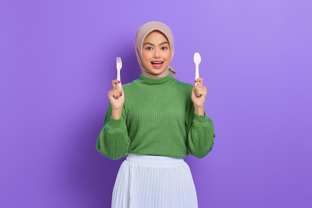 Cheerful beautiful asian woman in green sweater and hijab hold\
fork and spoon before breakfast isolated over purple\
background