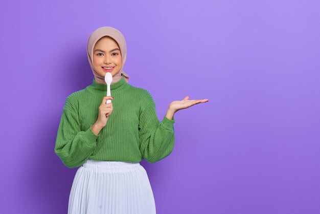 Cheerful beautiful Asian woman in green sweater and hijab holding a spoon and showing copy space on hands isolated over purple background