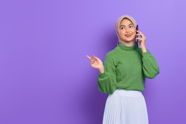 Cheerful beautiful Asian woman in green sweater and hijab talking on mobile phone while looking aside isolated over purple background