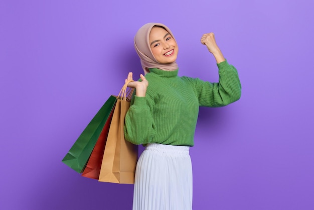 Cheerful beautiful asian woman in green sweater holding shopping bags and celebrate luck isolated over purple background