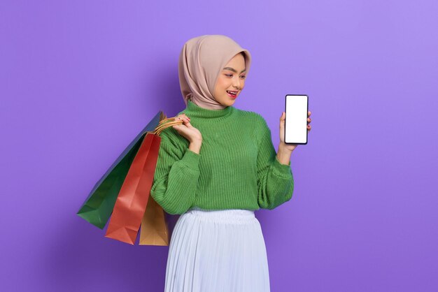 Cheerful beautiful asian woman in green sweater holding shopping bags, showing blank screen mobile phone isolated over purple background