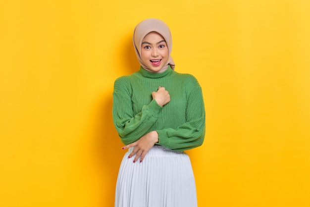 Cheerful beautiful Asian woman in green sweater pointing fingers at herself isolated over yellow background