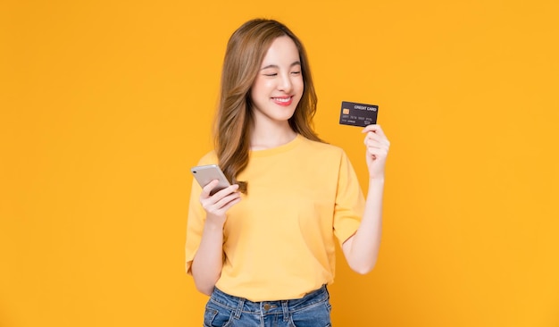 Cheerful beautiful Asian woman holding smartphone and mockup credit card on orange background