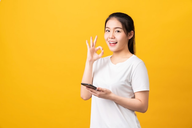 Cheerful beautiful Asian woman holding smartphone and shows ok sign