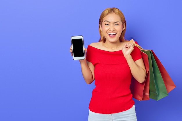 Cheerful beautiful asian woman in red dress holding shopping bags showing blank screen phone isolated over purple background