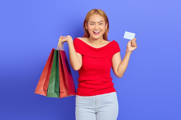 Cheerful beautiful asian woman in red dress holding shopping bags and showing credit card isolated over purple background