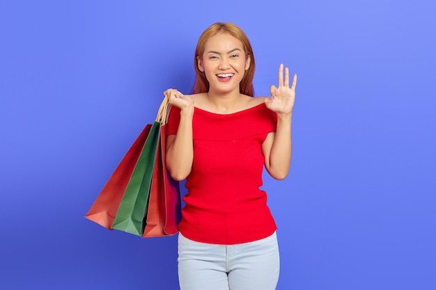 Cheerful beautiful Asian woman in red dress holding shopping bags, showing okay gesture isolated over purple background