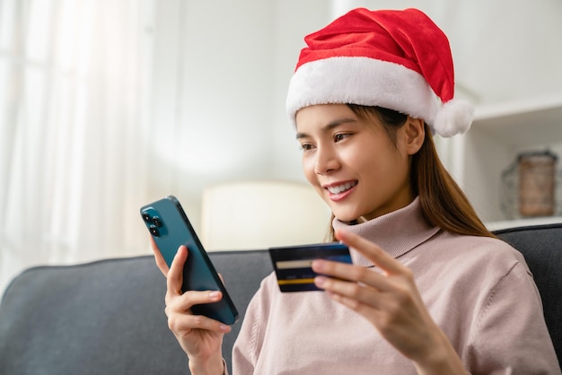 Foto allegra bella donna asiatica che indossa un cappello rosso di natale e tiene in mano una carta di credito e utilizza uno smartphone per lo shopping online con pagamento su internet banking