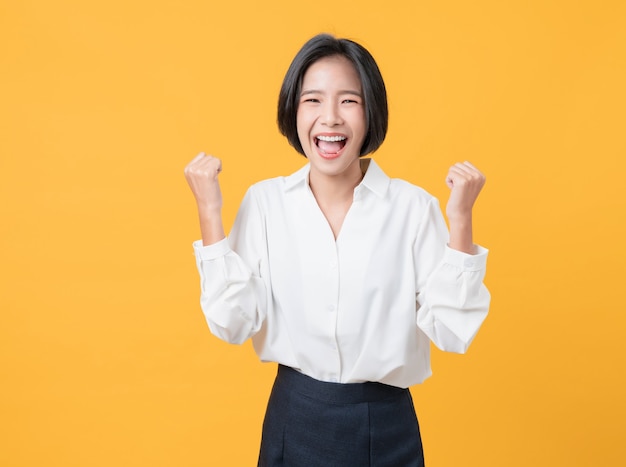 Cheerful beautiful Asian woman in a white shirt raises arms and fists clenched with shows strong powerful, celebrating victory expressing success.