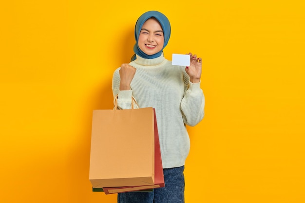 La bella donna asiatica allegra in maglione bianco che tiene la borsa della spesa e la carta di credito celebra la fortuna isolata sopra priorità bassa gialla