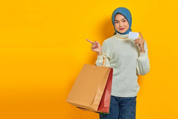 흰색 스웨터를 입은 쾌활한 아름다운 아시아 여성이 쇼핑백과 신용카드를 들고 노란색 배경에 격리된 손가락을 옆으로 가리키고 있습니다.