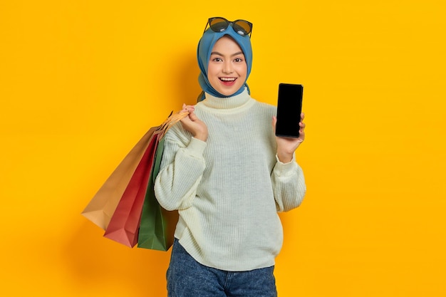 黄色の背景で隔離の空白の画面の携帯電話を示す買い物袋を保持している白いセーターで陽気な美しいアジアの女性