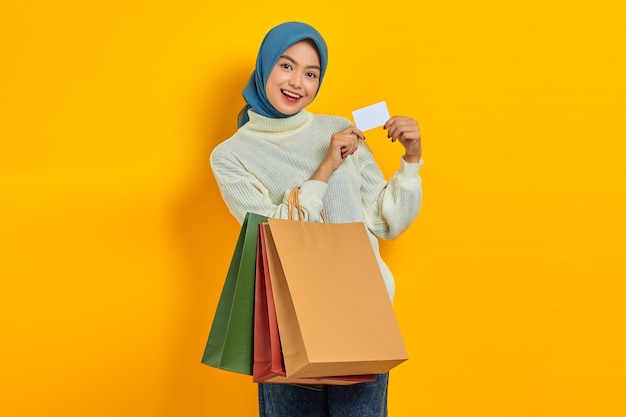 Cheerful beautiful Asian woman in white sweater holding shopping bags and showing credit card isolated over yellow background