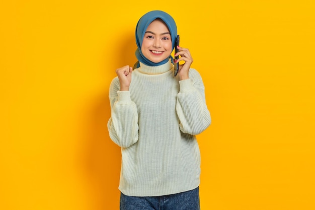 Cheerful beautiful Asian woman in white sweater talking mobile phone while rejoicing luck isolated over yellow background People islam religious concept