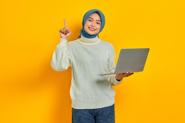 Cheerful beautiful Asian woman in white sweater using laptop and pointing fingers up having a good idea isolated over yellow background people religious lifestyle concept