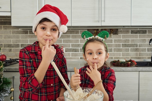 市松模様の赤と緑の服を着た元気で美しい子供たちが、クリスマスケーキのために用意した生地を味わいながら、口に指を持ってカメラを見ています。