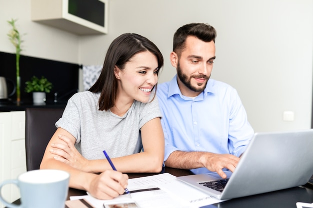 Cheerful beautiful couple sitting at table and using laptop while filling tax return form on website